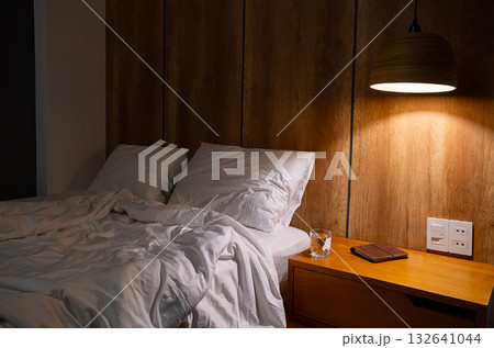 Unmade bed with white linen and pillows in hotel room with glass of water, book on nightstand at night. 132641044