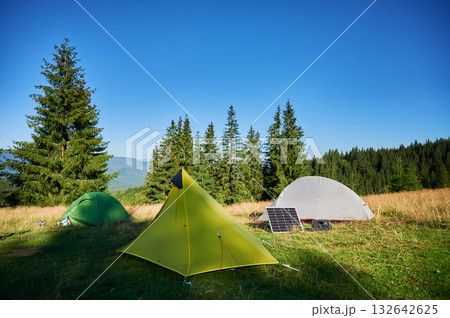 Tourist tent with solar panel and portable power station nearby in grassy field. Scene surrounded by rolling hills, tall pine trees under clear blue sky, showcasing eco-friendly camping setup. Tourist tent with solar panel and portable power station nearby in grassy field. Scene surrounded by rolling hills, tall pine trees under clear blue sky, showcasing eco-friendly camping setup. 132642625