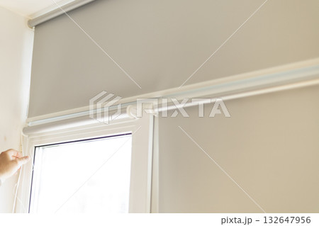 Process of installing light gray roller blinds on window indoors. 132647956