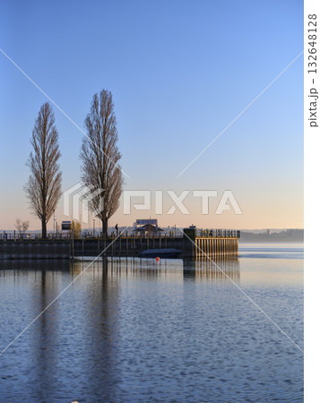 Serene morning at the lakeside dock with reflections and tall trees during sunrise 132648128