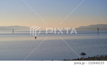 Calm waters of a lake reflecting the morning light with distant hills and markers along the shoreline Calm waters of a lake reflecting the morning light with distant hills and markers along the shoreline 132648155