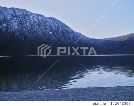 Serene view of a snow-capped mountain reflecting on calm lake waters at dusk near a pebble beach 132648398
