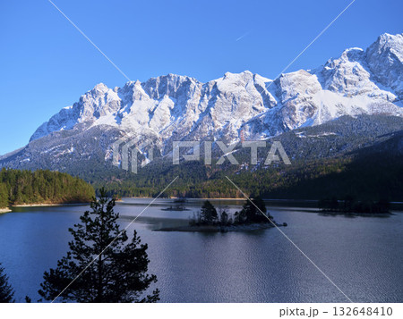 Snow-capped mountains reflect in calm lake surrounded by lush forest on a bright sunny day in the wilderness 132648410