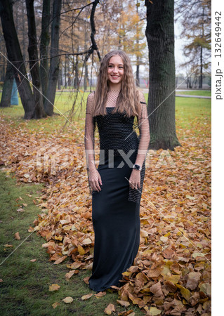 Young woman in a black mesh top stands among autumn trees and fallen leaves, looking away with a calm and thoughtful expression 132649442