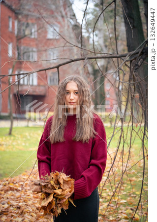 Teenage girl in a red sweater stands in a park holding autumn leaves, with soft light and warm tones creating a serene mood Teenage girl in a red sweater stands in a park holding autumn leaves, with soft light and warm tones creating a serene mood 132649447