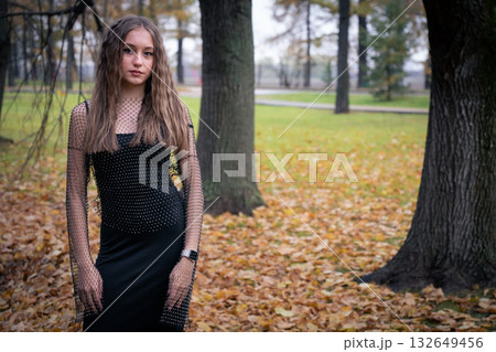 Young woman in a black mesh top stands among autumn trees and fallen leaves, looking away with a calm and thoughtful expression 132649456