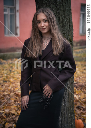 Young woman in a black mesh top and dark blazer stands near a tree with a muted red building in the background, looking away thoughtfully Young woman in a black mesh top and dark blazer stands near a tree with a muted red building in the background, looking away thoughtfully 132649458