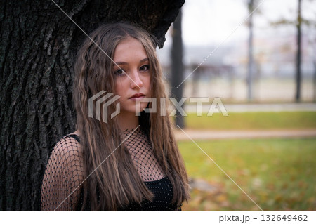 Teenage girl with long wavy hair in a black mesh top leans against a tree, surrounded by soft autumn light and muted tones 132649462