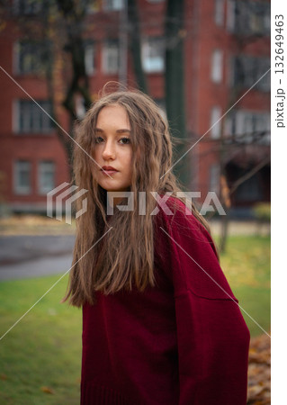 Teenage girl in a red sweater stands outdoors in soft autumn light, with blurred brick buildings and green grass in the background 132649463
