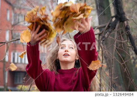Teenage girl in a red sweater joyfully throws autumn leaves into the air, smiling and looking up in a city park Teenage girl in a red sweater joyfully throws autumn leaves into the air, smiling and looking up in a city park 132649484