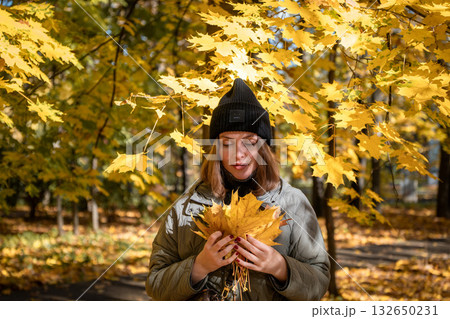 Young woman in a dark blazer and mesh top smiles softly while standing by a tree in an autumn park 132650231