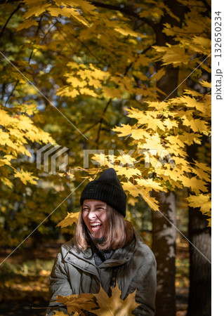 Woman in green jacket and black hat laughs under golden maple leaves in warm autumn sunlight Woman in green jacket and black hat laughs under golden maple leaves in warm autumn sunlight 132650234
