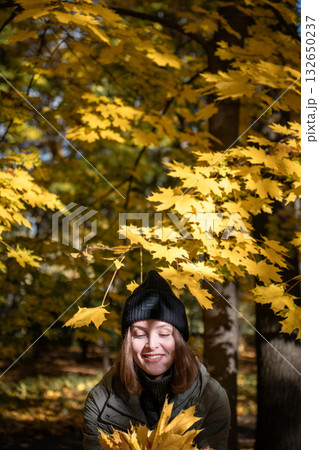 Woman in a green jacket and black hat smiles with eyes closed under golden maple leaves in warm autumn sunlight Woman in a green jacket and black hat smiles with eyes closed under golden maple leaves in warm autumn sunlight 132650237
