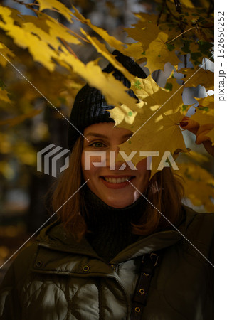 Woman in green jacket and knit hat smiles through a yellow maple leaf in sunlight among autumn trees 132650252