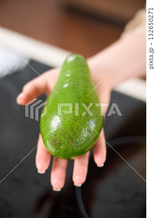 A large avocado in a woman's hand in the interiors of a bright and spacious kitchen 132650271