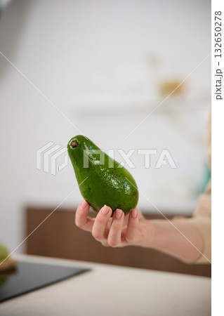 A large avocado in a woman's hand in the interiors of a bright and spacious kitchen 132650278