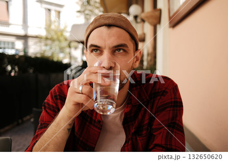 Young man in red checkered shirt and beanie drinking water at a cafe terrace Young man in red checkered shirt and beanie drinking water at a cafe terrace 132650620