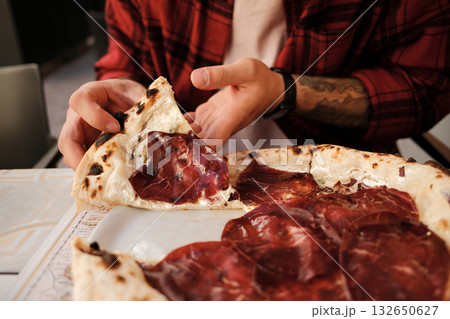 Close up of a man in a red checkered shirt enjoying pizza with cured meat at an outdoor cafe Close up of a man in a red checkered shirt enjoying pizza with cured meat at an outdoor cafe 132650627