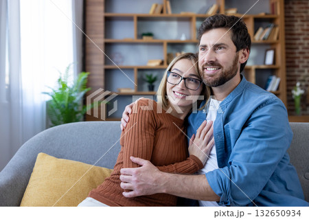 Loving couple sitting on a cozy sofa, hugging and smiling, looking away with happy expressions, embodying comfort, strong relationship, and a shared future inside a modern living room Loving couple sitting on a cozy sofa, hugging and smiling, looking away with happy expressions, embodying comfort, strong relationship, and a shared future inside a modern living room 132650934