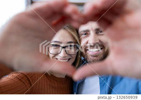 Smiling couple creating a heart with their hands and looking at the camera, conveying love, joy and close connection in a candid, cozy indoor portrait of partners 132650939