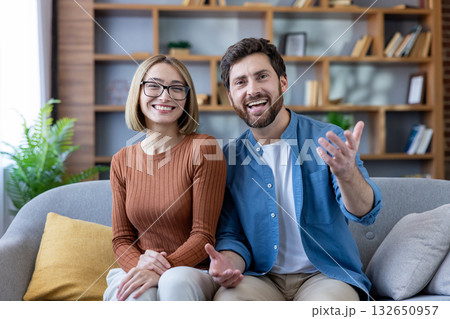 Cheerful young adult couple sitting on a cozy sofa, smiling and actively communicating during a virtual video conference, engaging in a pleasant remote conversation from their living room Cheerful young adult couple sitting on a cozy sofa, smiling and actively communicating during a virtual video conference, engaging in a pleasant remote conversation from their living room 132650957
