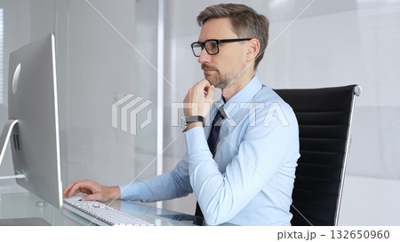 Businessman sitting at a clean office desk, wearing glasses and a light blue shirt with a tie, thinking and working on a computer, showing dedication and problem solving Businessman sitting at a clean office desk, wearing glasses and a light blue shirt with a tie, thinking and working on a computer, showing dedication and problem solving 132650960