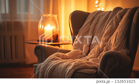 Cozy armchair draped with a knitted blanket, illuminated by warm candlelight, creating a serene atmosphere in a softly lit living room with gentle textures and inviting ambiance 132651025
