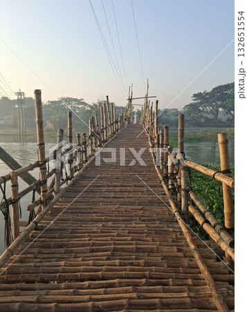 A rustic bamboo bridge stretches across a tranquil body of water, leading towards a distant, hazy horizon under a clear sky 132651554
