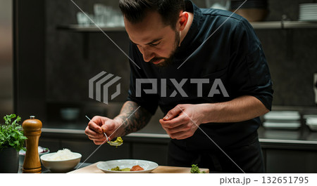 Male chef in black uniform meticulously plating gourmet dish in modern kitchen, showcasing culinary skills and attention to detail in a professional environment 132651795