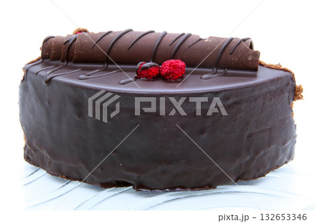 Close-up of a delicious slice of dark chocolate cake covered with glossy frosting. The dessert is decorated with chocolate curls and fresh raspberries, served on a transparent plate. Close-up of a delicious slice of dark chocolate cake covered with glossy frosting. The dessert is decorated with chocolate curls and fresh raspberries, served on a transparent plate. 132653346