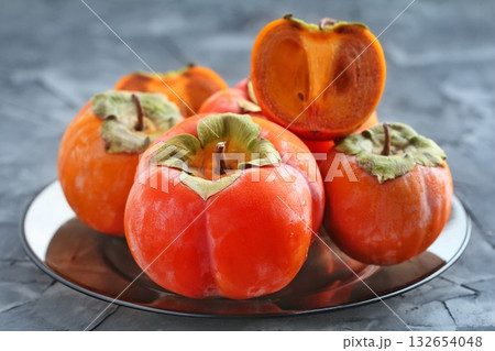 A group of vibrant orange persimmons, one sliced to reveal its rich interior, rests on a dark plate against a textured grey background. Concept of fresh, nutritious fruit. 132654048