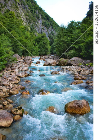A vibrant blue-green river rushes over numerous rocks through a lush green valley with steep, rocky mountainsides. Concept of majestic wilderness and powerful nature. 132654166