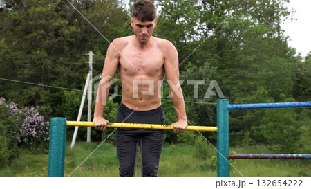 Hardy shirtless sportsman doing push ups on horizontal bar at nature. Strong and muscular guy training at sports ground. Young athletic guy exercising outdoor. Concept of active and healthy lifestyle 132654222