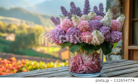 Glass vase with vibrant arrangement of pink and white hydrangea flowers, filled with purple cabbage leaves at bottom, placed outdoors with golden hour light in background. 132654774