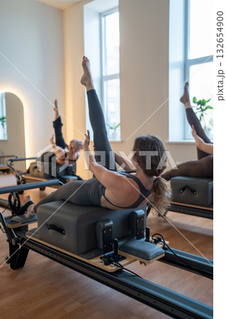 Group of caucasian women exercising with trainer on pilates reformers in a bright studio. Their workout focuses on core strength and flexibility in a modern fitness environment Group of caucasian women exercising with trainer on pilates reformers in a bright studio. Their workout focuses on core strength and flexibility in a modern fitness environment 132654900
