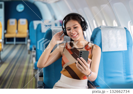 Tourist and Inflight Entertainment. A woman with headphones smiling while using her smartphone on an airplane. 132662734