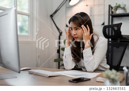 Stressed Asian female call center agent managing customer inquiries at a modern workspace, showcasing the challenges of remote work. 132663581