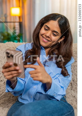 Indian woman feeling nostalgic while looking at old photos on smartphone at home, sits on soft sofa 132666553
