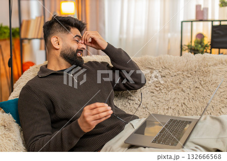 Indian man rubs tired eyes while on sofa with laptop at home, eyes strained after long screen time Indian man rubs tired eyes while on sofa with laptop at home, eyes strained after long screen time 132666568
