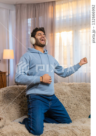 Young man dancing on couch with wireless headphones playing air guitar and enjoying disco music 132666992