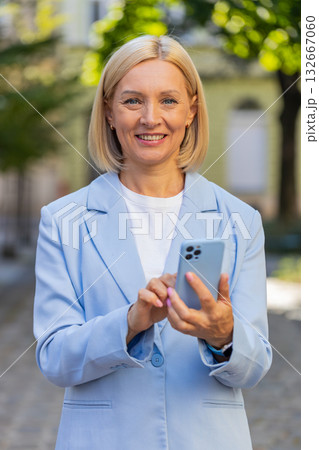 Smiling mature Caucasian businesswoman in suit using smartphone and looking at camera on city street Smiling mature Caucasian businesswoman in suit using smartphone and looking at camera on city street 132667060