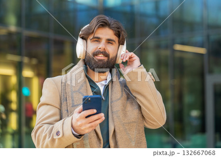 Happy young man putting on headphones listening favorite energetic music on smartphone on street Happy young man putting on headphones listening favorite energetic music on smartphone on street 132667068