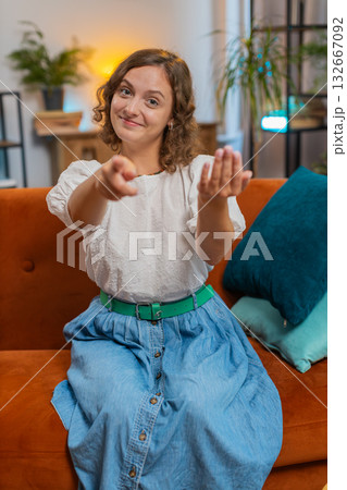 Young woman smiling and pointing to camera, choosing inviting, select sitting on sofa couch at home 132667092