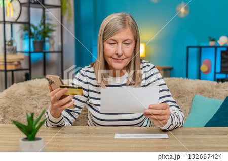 Mature woman using credit card and smartphone to transfer money, pay bills, sitting on table at home Mature woman using credit card and smartphone to transfer money, pay bills, sitting on table at home 132667424