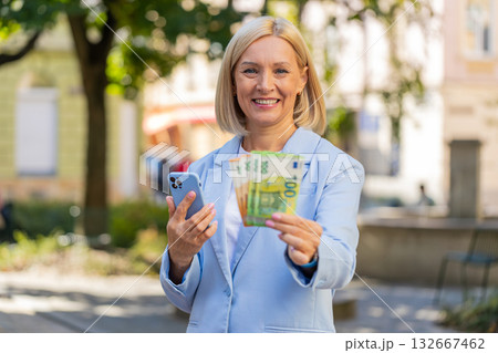 Caucasian mature businesswoman on city street showing earnings cash money with smartphone in hands Caucasian mature businesswoman on city street showing earnings cash money with smartphone in hands 132667462