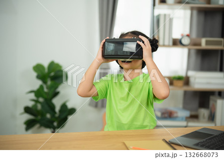 Child Engaged in Virtual Reality Experience with VR Headset while Sitting at Table in Modern Room with Laptop and Indoor Plant in Background Child Engaged in Virtual Reality Experience with VR Headset while Sitting at Table in Modern Room with Laptop and Indoor Plant in Background 132668073