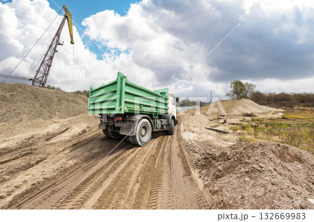 A sturdy green truck moves along a rugged dirt path beside a construction area, surrounded by mounds of earth and a crane in the distance on a cloudy day 132669983