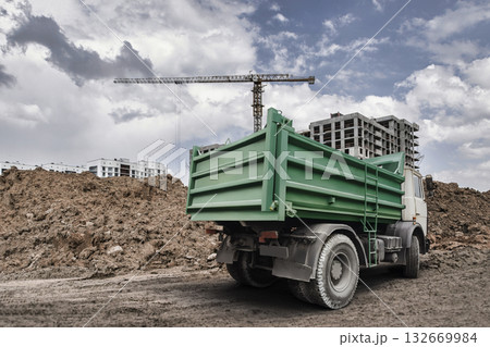 Bright green dump truck sits on a construction site with piles of dirt, cranes in the distance, and clouds creating an impressive backdrop 132669984