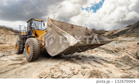 A large excavator loader digs into the sandy terrain at a construction site, surrounded by clouds softly drifting across the blue sky. 132670004