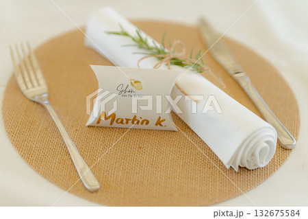 Elegant table setting features a personalized name card, silver cutlery, and a rolled white napkin adorned with rosemary on a woven placemat, creating an inviting dining atmosphere 132675584